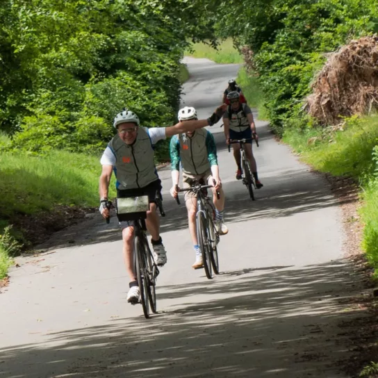 Three people on their bikes cycling along a leafy lane. Man at the front looks happy with his arm out